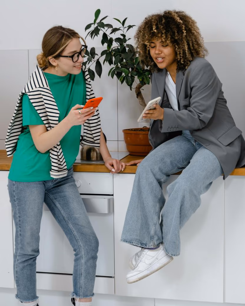 Two housemates in a kitchen, comparing something on their phones.
